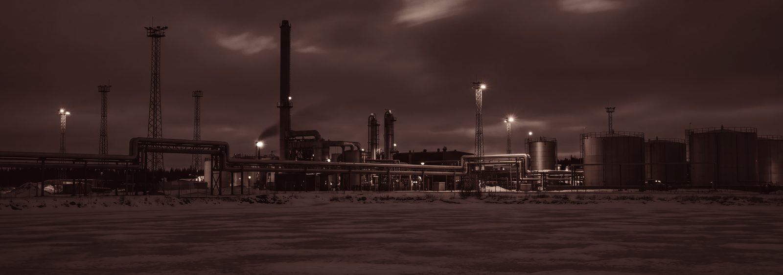 Industriell anläggning i vinternatt: pipor, cisterner och arbetsbelysning över snötäckt mark. Inga människor och inga fordon.